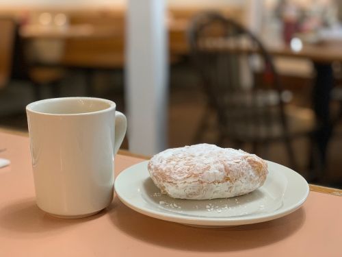 A mug of coffee sits on a table next to a jelly filled donut covered in sugar which is sitting on a plate